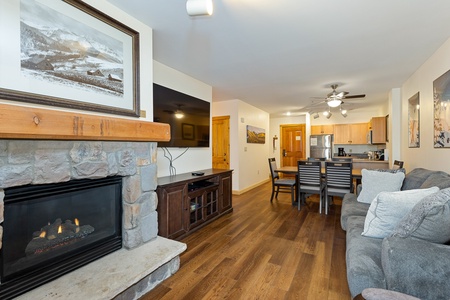 A cozy living room with a stone fireplace, TV on a wooden stand, gray sofa, and dining area. The kitchen is in the background with modern appliances. Framed pictures decorate the white walls.