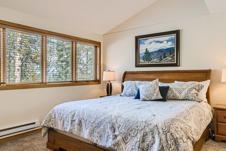 A bedroom with a large bed featuring patterned bedspread and pillows, flanked by two bedside tables with lamps. A framed landscape painting hangs above the bed, and a window with blinds is on the left.