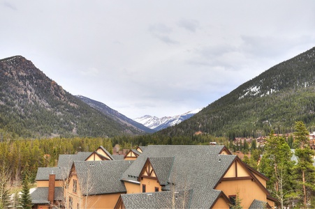 Row of brown houses with dark roofs nestled in a valley surrounded by green, forested mountains with snow-covered peaks in the distance under a cloudy sky.