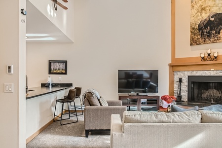 A living room with a beige couch, a gray armchair, a TV on a stand, a fireplace with a stone surround, and a kitchen area with a counter and two bar stools. A painting hangs above the fireplace.