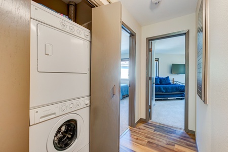 A small laundry area with a stacked washer and dryer is located in a hallway. The hallway leads to a bedroom with a blue bedspread and a wall-mounted TV.