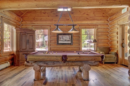 A log cabin room with wooden walls and floor, featuring a pool table in the center. There is an armoire on the left, windows behind the table, and a chair to the right near a door.