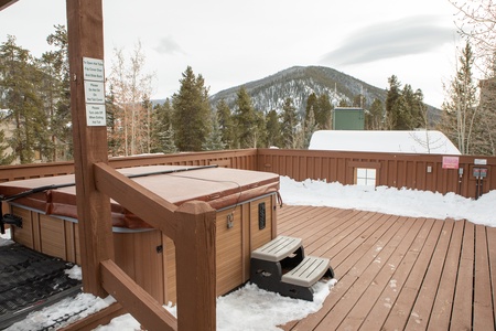 Outdoor hot tub on a wooden deck in a snowy mountain setting, surrounded by trees and a snow-covered roof in the background.