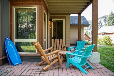 A patio with wooden and blue plastic chairs, set against a house with large windows and a sliding door. A blue sled is propped against the wall. Trees and another building are visible in the background.