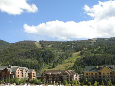 A row of buildings sits at the base of a green, tree-covered mountain with ski lifts visible, under a sky with clouds.