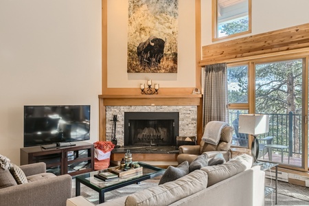 A living room featuring a fireplace with a large bison artwork above it, cozy seating, a television, and a glimpse of a balcony with trees outside.