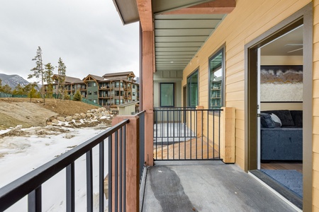 A balcony with mountain views extends from a modern building. It features a doorway leading to a room with a blue sofa and a large painting on the wall. Snow-covered ground is visible below.