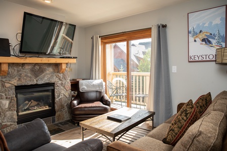 A cozy living room with a stone fireplace, mounted TV, and a brown armchair. There's a couch, coffee table, and a sliding glass door leading to a balcony. A "Keystone" poster is on the wall.