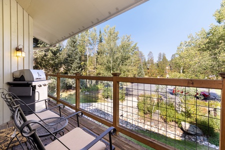 A balcony with two chairs and a barbecue grill overlooks a parking area and trees under a clear sky.