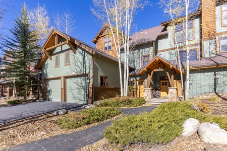 A two-story house with green siding, wooden accents, and a two-car garage, surrounded by trees and shrubs on a clear, sunny day.