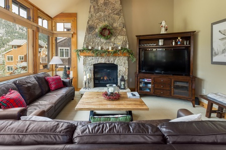 A cozy living room features large windows, a stone fireplace with a wreath, two brown leather couches, a wooden coffee table, and a TV on a wooden stand.