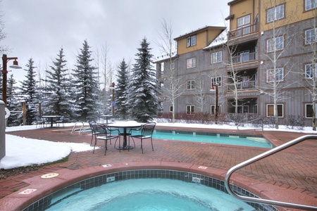 Outdoor pool and hot tub area of a residential building, surrounded by snow-covered trees and ground. The poolside has metal chairs and tables, and the sky is overcast.