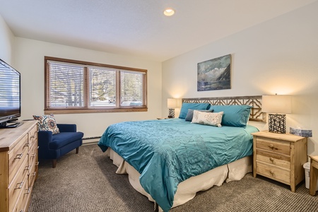 A bedroom with a large bed featuring turquoise bedding, two bedside tables with lamps, a dresser with a TV, and a window with wooden blinds. A chair is placed next to the window.