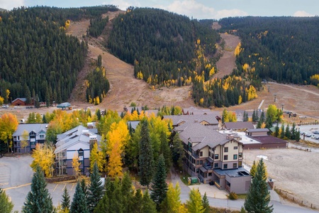 A resort-style complex with multiple buildings is surrounded by dense forests and hills, featuring ski runs on the slopes in the background, covered in autumn foliage.