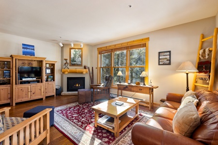 A cozy living room features a leather sofa, wooden furniture, a fireplace, a large window with natural light, and a red patterned rug.