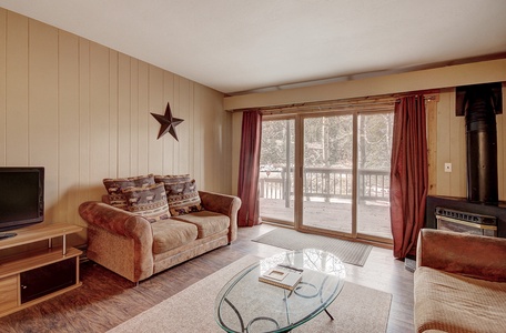 A cozy living room with wooden paneling, a couch, a TV on a stand, a glass coffee table, and sliding glass doors leading to a deck.