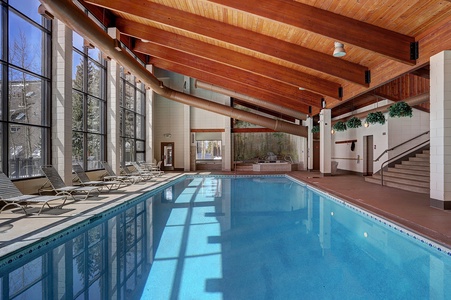 Indoor swimming pool with chairs lined on the deck, large windows allowing natural light, wooden ceiling with beams, spiral slide, and stairs leading to an upper level.