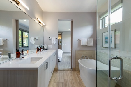 Modern bathroom with double vanity, wall mirrors, and under-counter storage. Features a white bathtub near a glass-enclosed shower, white walls, and light wood flooring. Bedroom visible in the background.