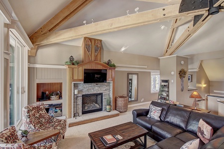 A cozy living room with exposed wooden beams, a stone fireplace, a black leather sofa, patterned chairs, and a wooden coffee table. The space features neutral tones and large windows.