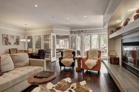 A living room with a beige couch, two armchairs with fur throws, a round wooden coffee table, and a fireplace. Dining area with a chandelier and glass doors leading to an outdoor patio.