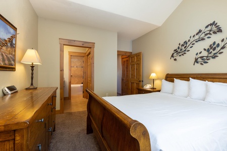 A cozy bedroom with a wooden bed frame, white bedding, two nightstands with lamps, a wooden dresser, and wall-mounted metal leaf decor. An open doorway reveals a bathroom in the background.