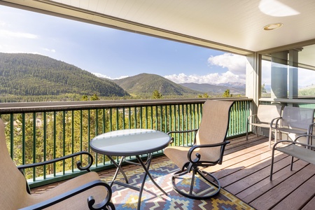 A covered balcony with two chairs, a small round table, and a patterned rug offers a view of a forested mountain landscape under a clear sky.