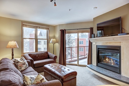 A cozy living room with two brown sofas, a wooden coffee table, a fireplace, a TV above the fireplace, and a sliding glass door leading to a snowy balcony. Lamps flank one of the sofas.