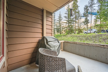 A small covered patio with a grill, wicker chairs, and a view of a grassy area with trees and a pickup truck in the background.