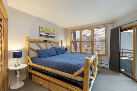 A bedroom with a wooden bed frame, blue bedding, a nightstand with a lamp, a painting above the bed, and large windows allowing natural light. There is also a partially open door to a balcony.