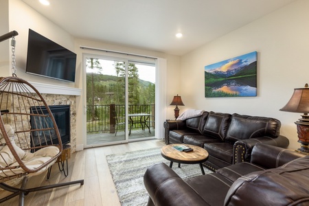 A modern living room with a hanging chair, black leather couches, a coffee table, a fireplace, wall art, and a TV. A sliding glass door leads to a balcony with a scenic view outside.