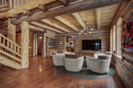 A log cabin living room with wooden beams and floor, modern furniture including a circular arrangement of four armchairs, a large TV, a contemporary light fixture, and a staircase leading upstairs.