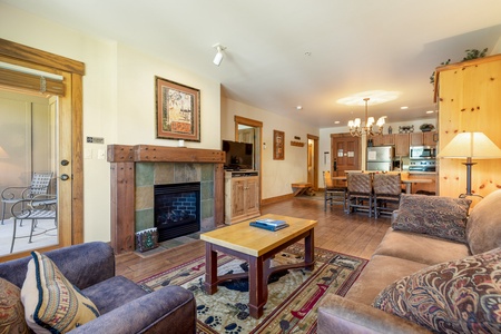 A cozy living room with a fireplace, wooden furniture, a couch, armchair, and a TV. A dining table is in the background next to a kitchen with stainless steel appliances. A patio is visible through a door.