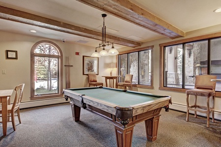 A game room with a pool table in the center, a table with chairs on the left, bar stools along the walls, large windows, and a wooden beam ceiling.