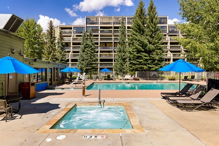 Outdoor pool area featuring a large pool and a hot tub with several blue umbrellas and lounge chairs. A multi-story building with balconies is in the background, surrounded by tall trees.