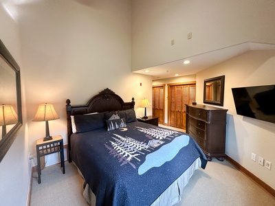 A bedroom with a wooden bed, tree-patterned bedspread, two nightstands with lamps, a dresser, and a wall-mounted TV. The room has carpeted flooring and a wooden closet.