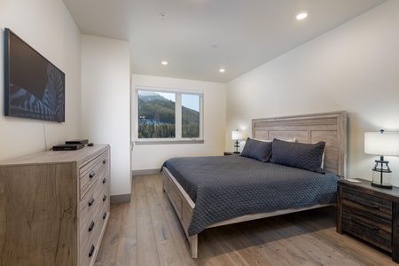 A modern bedroom with a queen-sized bed, two bedside tables with lamps, a TV mounted on the wall, and a dresser. A window offers a view of a forested hillside.