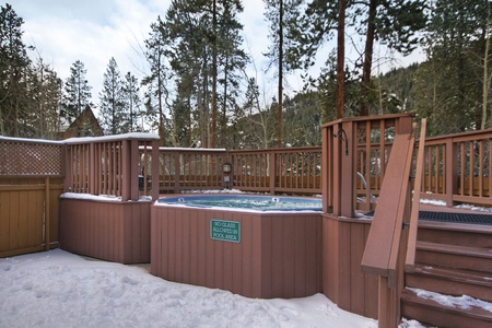 An outdoor hot tub surrounded by a wooden deck and railing, with snow-covered ground and trees in the background. A sign on the hot tub says, "No Glass Allowed In Pool Area.