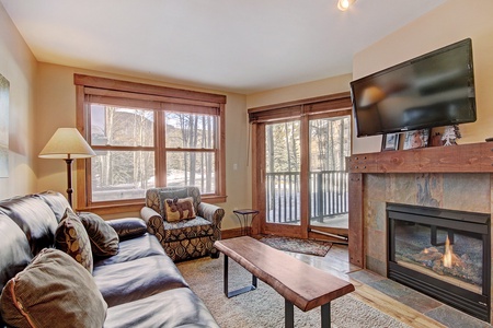 A cozy living room with a leather sofa, armchair, wood coffee table, fireplace, and wall-mounted TV. Large windows and glass doors reveal a snowy outdoor scene.