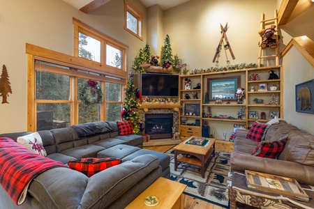 A cozy living room with a fireplace, decorated for Christmas with a tree, stockings, and holiday ornaments. Two large sofas, a coffee table, and shelves with various items surround the space.