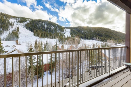 A snow-covered mountain landscape is viewed from a balcony with a railing, trees, and buildings partially visible below. The sky is partly cloudy.