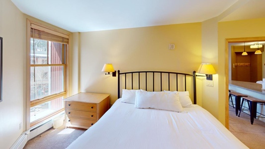 A bedroom with a double bed, white bedding, wooden side table, wall-mounted lamps, and a window with wooden blinds. A doorway leads to another room with bar stools and pendant lights.