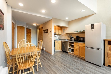 A kitchen with wooden cabinets and stainless steel appliances, including a refrigerator, dishwasher, and microwave. A wooden dining table with four chairs is in the foreground.