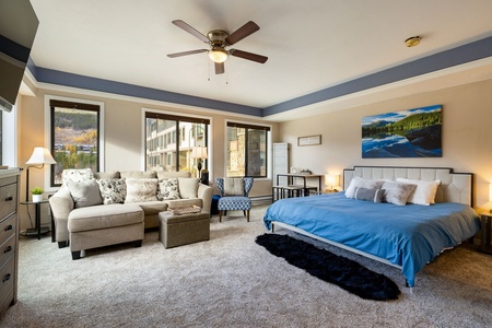 A spacious bedroom with a ceiling fan, a king-sized bed with blue bedding, a beige sectional sofa, a black rug, wall art, and large windows.