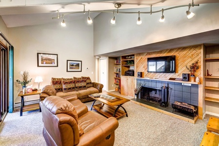 A cozy living room with leather chairs, a patterned sofa, a coffee table, and a fireplace with a mounted TV. Shelves flank the fireplace, and a track light fixture is installed on the ceiling.