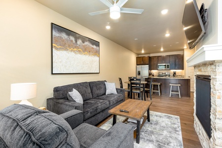 Modern living room and kitchen area featuring gray furniture, a wooden coffee table, wall art, ceiling fan, and a mounted TV above a fireplace. The kitchen has dark wood cabinets and a breakfast bar.