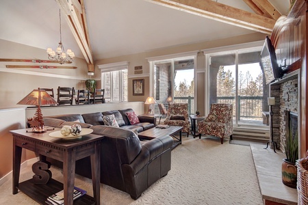 A living room with a sectional sofa, coffee table, decorative lamp, two chairs, a dining table, wooden ceiling beams, and a mounted TV next to a stone fireplace. Large windows show an outdoor view.