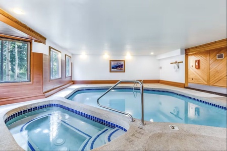 A spacious indoor pool area featuring a hot tub on the left, large windows along one wall, a painting on the far wall, wooden paneling, and a handrail leading into the pool.