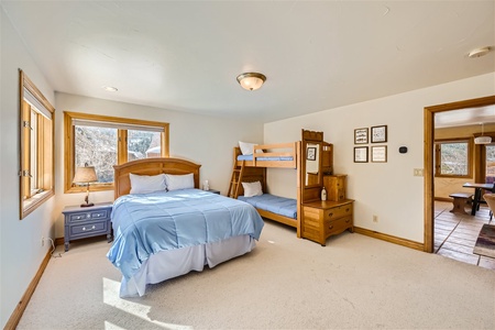 A bedroom with a queen bed, bunk bed, and wooden furniture. It has two windows, beige carpeting, and an adjoining doorway leading to another room.