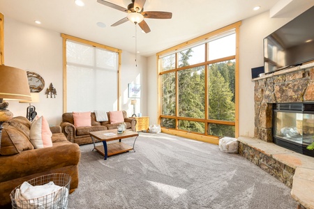 A cozy living room with large windows, a ceiling fan, a brown sofa, a stone fireplace, and a TV mounted above. The room is well-lit with natural light and has a view of tall trees outside.