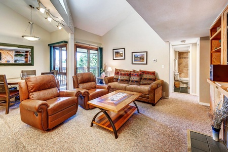 A living room with two brown leather recliners, a beige sofa, a wooden coffee table, a dining table, and large glass doors leading to a balcony. The background shows a hallway and a bathroom.